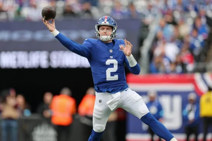 Drew Lock in einer blauen Uniform mit der Nummer 2, die an die Farben der New York Giants erinnert, wirft einen Pass auf das Spielfeld. Die dynamische Szene fängt seinen ausgestreckten Arm ein, während der Ball durch die Luft segelt, vor einem Hintergrund aus verschwommenen Zuschauern und Stadionbeschilderung. Diese Beschreibung wurde mit der FootballR KI automatisch generiert.