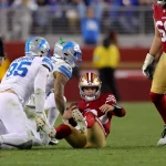Brock Purdy in rot-goldener Uniform sitzt auf dem Feld und blickt nach oben. Zwei gegnerische Spieler in weiß-blauen Uniformen knien in der Nähe. Im Hintergrund steht ein Schiedsrichter in schwarz-weißen Streifen mit erhobenem Arm. In der Nähe überblickt NFL-Aufsteiger Brock Purdy die Szene von rechts. Diese Beschreibung wurde mit der FootballR KI automatisch generiert.