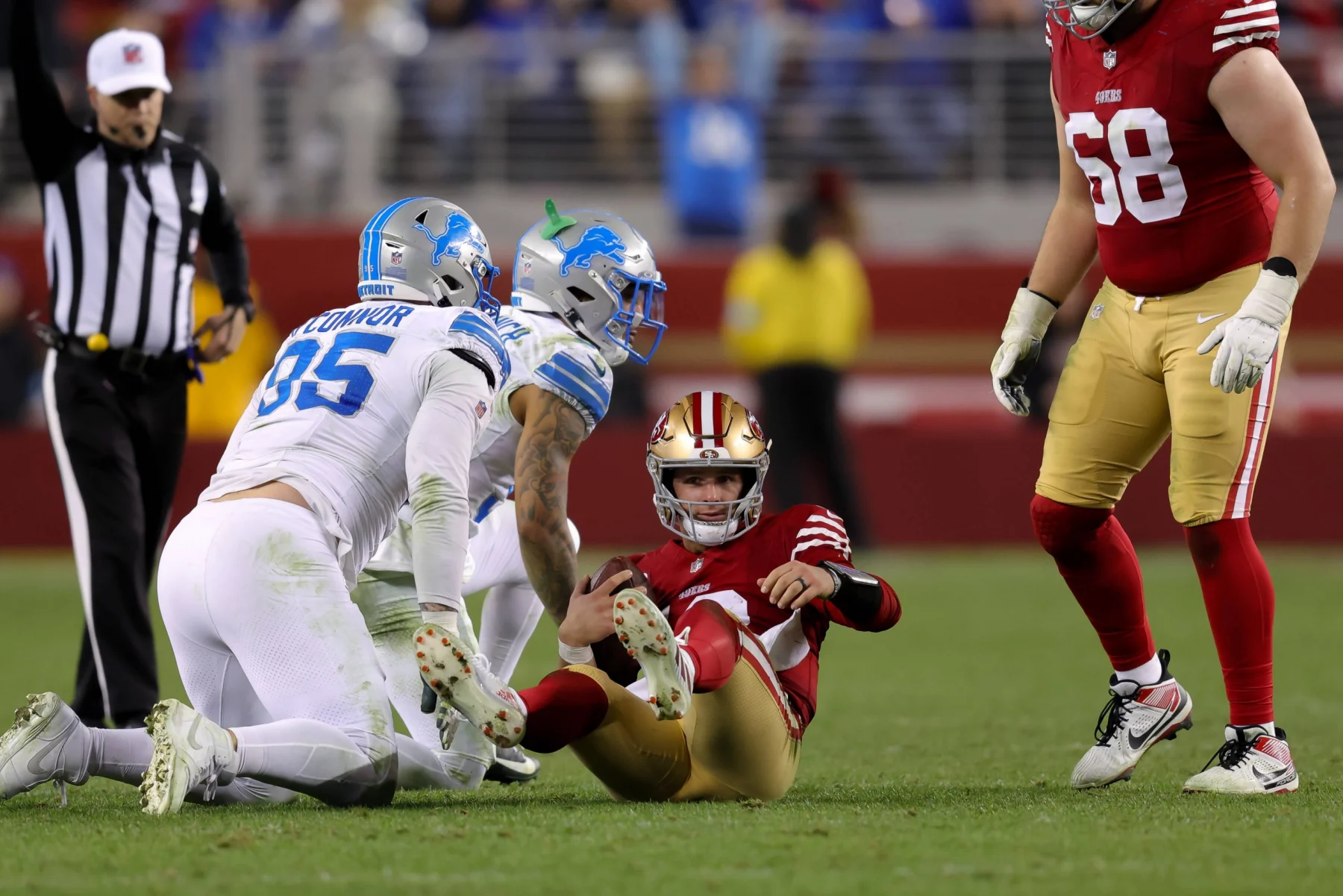 Brock Purdy in rot-goldener Uniform sitzt auf dem Feld und blickt nach oben. Zwei gegnerische Spieler in weiß-blauen Uniformen knien in der Nähe. Im Hintergrund steht ein Schiedsrichter in schwarz-weißen Streifen mit erhobenem Arm. In der Nähe überblickt NFL-Aufsteiger Brock Purdy die Szene von rechts. Diese Beschreibung wurde mit der FootballR KI automatisch generiert.