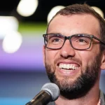 Andrew Luck mit kurzen Haaren und Brille, der an Andrew Luck erinnert, lächelt, während er in ein Mikrofon spricht. Der Hintergrund ist mit weichen, bunten Lichtern verschwommen, wodurch ein Bokeh-Effekt entsteht. Er trägt eine dunkle Jacke über einem hellen Hemd. Diese Beschreibung wurde mit der FootballR KI automatisch generiert.