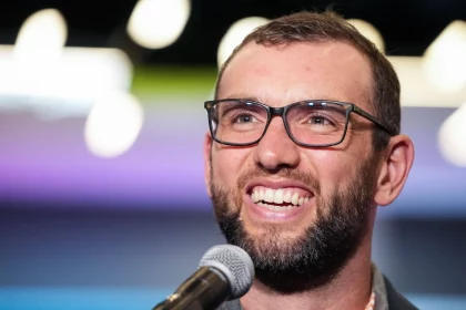 Andrew Luck mit kurzen Haaren und Brille, der an Andrew Luck erinnert, lächelt, während er in ein Mikrofon spricht. Der Hintergrund ist mit weichen, bunten Lichtern verschwommen, wodurch ein Bokeh-Effekt entsteht. Er trägt eine dunkle Jacke über einem hellen Hemd. Diese Beschreibung wurde mit der FootballR KI automatisch generiert.