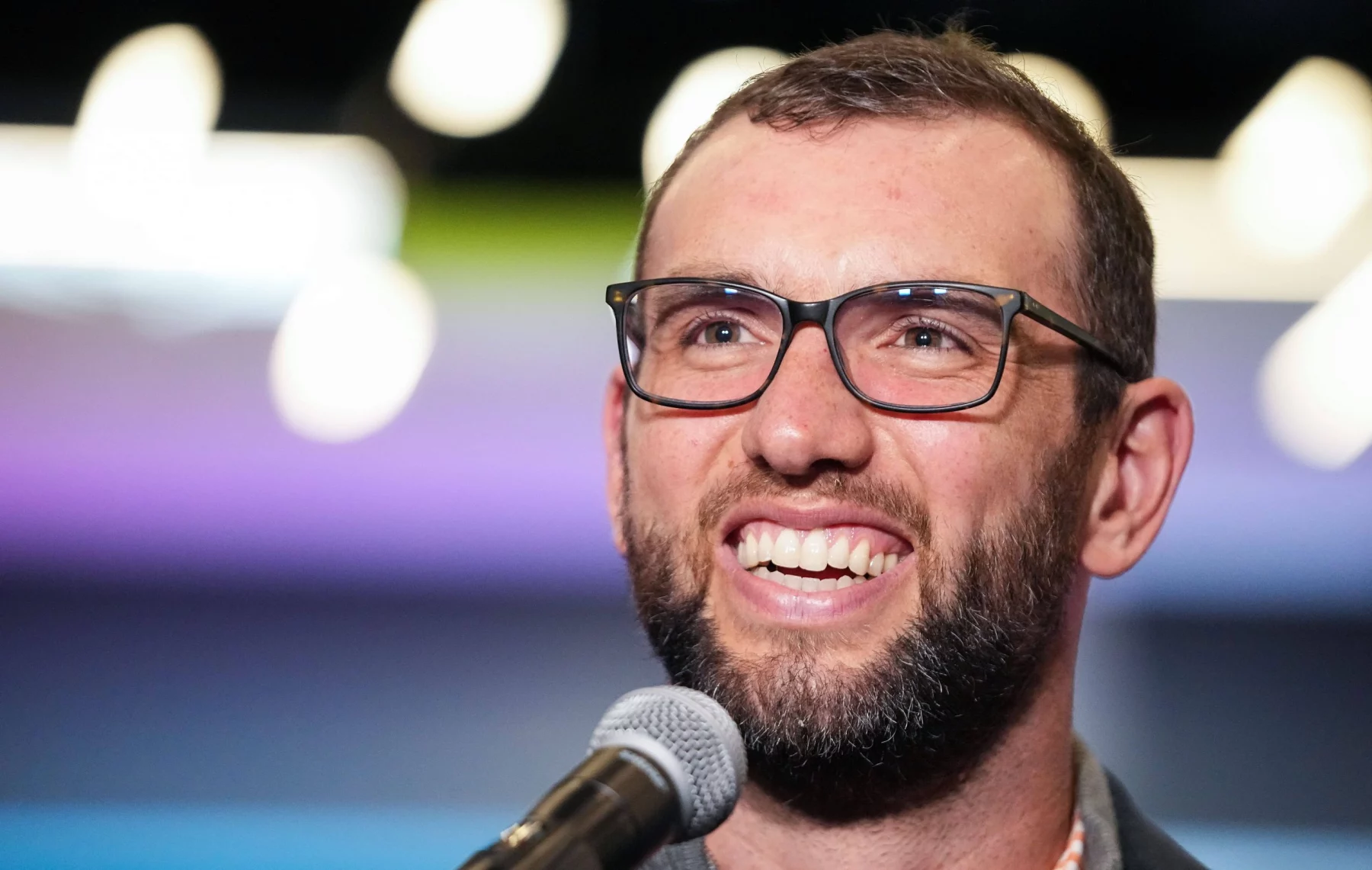 Andrew Luck mit kurzen Haaren und Brille, der an Andrew Luck erinnert, lächelt, während er in ein Mikrofon spricht. Der Hintergrund ist mit weichen, bunten Lichtern verschwommen, wodurch ein Bokeh-Effekt entsteht. Er trägt eine dunkle Jacke über einem hellen Hemd. Diese Beschreibung wurde mit der FootballR KI automatisch generiert.