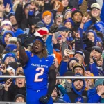 Ashton Jeanty in einem blauen Trikot mit der Nummer 2 feiert mit einer jubelnden Menge in Winterkleidung. Fans, die von den jüngsten College Football Awards begeistert sind, halten ihre Handys hoch, um den Moment festzuhalten. Die festliche Atmosphäre ist geprägt von farbenfroher Teamausrüstung und begeisterten Gesichtsausdrücken. Diese Beschreibung wurde mit der FootballR KI automatisch generiert.