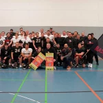 Eine große Gruppe von Männern, einige in American-Football-Ausrüstung, andere in Freizeitkleidung, posiert drinnen auf einem Sportplatz. Sie halten Fanartikel ihrer Mannschaft in den Händen, darunter eine große Flagge mit „Lüneburg Razorbacks“ und einem Wildschwein-Logo. Helme liegen vor ihnen auf dem Boden. Lächelnde Gesichter und ein Gemeinschaftsgefühl beim American Football sind deutlich zu erkennen. Diese Beschreibung wurde mit der FootballR KI automatisch generiert.