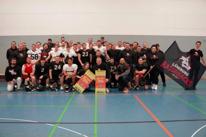 Eine große Gruppe von Männern, einige in American-Football-Ausrüstung, andere in Freizeitkleidung, posiert drinnen auf einem Sportplatz. Sie halten Fanartikel ihrer Mannschaft in den Händen, darunter eine große Flagge mit „Lüneburg Razorbacks“ und einem Wildschwein-Logo. Helme liegen vor ihnen auf dem Boden. Lächelnde Gesichter und ein Gemeinschaftsgefühl beim American Football sind deutlich zu erkennen. Diese Beschreibung wurde mit der FootballR KI automatisch generiert.