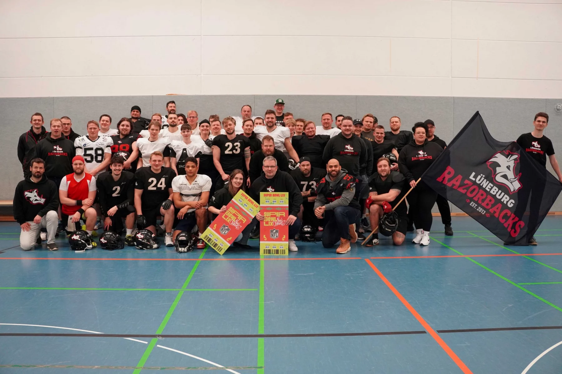 Eine große Gruppe von Männern, einige in American-Football-Ausrüstung, andere in Freizeitkleidung, posiert drinnen auf einem Sportplatz. Sie halten Fanartikel ihrer Mannschaft in den Händen, darunter eine große Flagge mit „Lüneburg Razorbacks“ und einem Wildschwein-Logo. Helme liegen vor ihnen auf dem Boden. Lächelnde Gesichter und ein Gemeinschaftsgefühl beim American Football sind deutlich zu erkennen. Diese Beschreibung wurde mit der FootballR KI automatisch generiert.