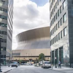 New Orleans - Caesars Superdome: Eine breite Straßenansicht einer modernen Stadtlandschaft mit hohen Gebäuden umrahmt eine große, einzigartig geschwungene Struktur im Hintergrund unter einem bewölkten Himmel. Fahrzeuge und Fußgänger, darunter Football-Fans, die sich auf den Super Bowl LIX freuen, füllen den Vordergrund, wobei das geschwungene Gebäude den Mittelpunkt bildet. Diese Beschreibung wurde mit der FootballR KI automatisch generiert.