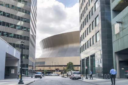 New Orleans - Caesars Superdome: Eine breite Straßenansicht einer modernen Stadtlandschaft mit hohen Gebäuden umrahmt eine große, einzigartig geschwungene Struktur im Hintergrund unter einem bewölkten Himmel. Fahrzeuge und Fußgänger, darunter Football-Fans, die sich auf den Super Bowl LIX freuen, füllen den Vordergrund, wobei das geschwungene Gebäude den Mittelpunkt bildet. Diese Beschreibung wurde mit der FootballR KI automatisch generiert.