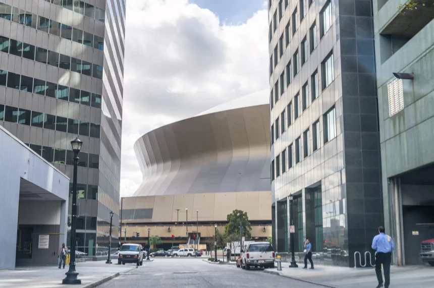 New Orleans - Caesars Superdome: Eine breite Straßenansicht einer modernen Stadtlandschaft mit hohen Gebäuden umrahmt eine große, einzigartig geschwungene Struktur im Hintergrund unter einem bewölkten Himmel. Fahrzeuge und Fußgänger, darunter Football-Fans, die sich auf den Super Bowl LIX freuen, füllen den Vordergrund, wobei das geschwungene Gebäude den Mittelpunkt bildet. Diese Beschreibung wurde mit der FootballR KI automatisch generiert.