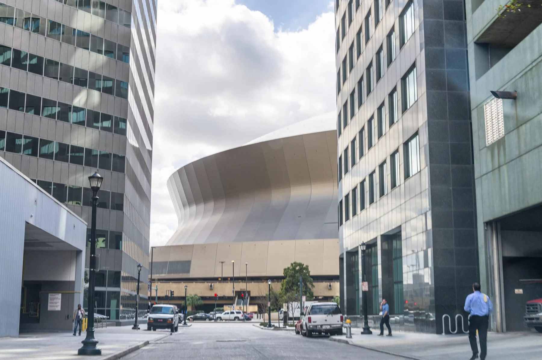 New Orleans - Caesars Superdome: Eine breite Straßenansicht einer modernen Stadtlandschaft mit hohen Gebäuden umrahmt eine große, einzigartig geschwungene Struktur im Hintergrund unter einem bewölkten Himmel. Fahrzeuge und Fußgänger, darunter Football-Fans, die sich auf den Super Bowl LIX freuen, füllen den Vordergrund, wobei das geschwungene Gebäude den Mittelpunkt bildet. Diese Beschreibung wurde mit der FootballR KI automatisch generiert.