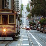 Ein klassisches Cable Car aus San Francisco mit einem Exklusiver-Sponsor-Schild fährt eine steile Straße hinauf, die von parkenden Autos, Gebäuden und Bäumen gesäumt ist. Auf der Seilbahn steht "Van Ness Ave, California & Market Streets", während vor ihr eine Ampel leuchtet. Diese Beschreibung wurde mit der FootballR KI automatisch generiert.
