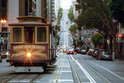 Ein klassisches Cable Car aus San Francisco mit einem Exklusiver-Sponsor-Schild fährt eine steile Straße hinauf, die von parkenden Autos, Gebäuden und Bäumen gesäumt ist. Auf der Seilbahn steht "Van Ness Ave, California & Market Streets", während vor ihr eine Ampel leuchtet. Diese Beschreibung wurde mit der FootballR KI automatisch generiert.