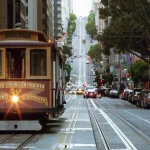 Ein klassisches Cable Car aus San Francisco mit einem Exklusiver-Sponsor-Schild fährt eine steile Straße hinauf, die von parkenden Autos, Gebäuden und Bäumen gesäumt ist. Auf der Seilbahn steht "Van Ness Ave, California & Market Streets", während vor ihr eine Ampel leuchtet. Diese Beschreibung wurde mit der FootballR KI automatisch generiert.