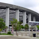 Eingang zum SoFi-Stadion mit einem großen Schild mit der Aufschrift „SoFi-Stadion“ auf dem gewölbten Dach. Im Vordergrund zeigt Brown Lot D markierte Eingänge und eine Karte, die die Fans der Los Angeles Rams willkommen heißt. Unter einem bewölkten Himmel sind mehrere Eingangsschilder, Tore und Grünflächen zu sehen. Diese Beschreibung wurde mit der FootballR KI automatisch generiert.