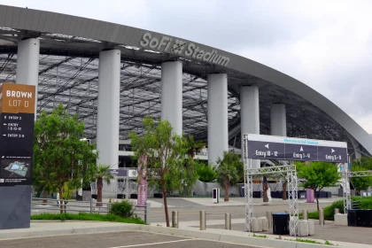 Eingang zum SoFi-Stadion mit einem großen Schild mit der Aufschrift „SoFi-Stadion“ auf dem gewölbten Dach. Im Vordergrund zeigt Brown Lot D markierte Eingänge und eine Karte, die die Fans der Los Angeles Rams willkommen heißt. Unter einem bewölkten Himmel sind mehrere Eingangsschilder, Tore und Grünflächen zu sehen. Diese Beschreibung wurde mit der FootballR KI automatisch generiert.