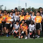 Eine Gruppe junger Sportler in orange-weißen Uniformen posiert auf einem Rasenplatz und hält einen Football in der Hand, als wären sie bereit für den Pro Bowl. Sie sind von Trainern und Schiedsrichtern in schwarz-weiß gestreiften Hemden umgeben. Der Himmel ist klar, Bäume rahmen die Szene ein und alle sehen glücklich und aufgeregt aus. Diese Beschreibung wurde mit der FootballR KI automatisch generiert.