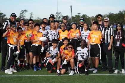 Eine Gruppe junger Sportler in orange-weißen Uniformen posiert auf einem Rasenplatz und hält einen Football in der Hand, als wären sie bereit für den Pro Bowl. Sie sind von Trainern und Schiedsrichtern in schwarz-weiß gestreiften Hemden umgeben. Der Himmel ist klar, Bäume rahmen die Szene ein und alle sehen glücklich und aufgeregt aus. Diese Beschreibung wurde mit der FootballR KI automatisch generiert.
