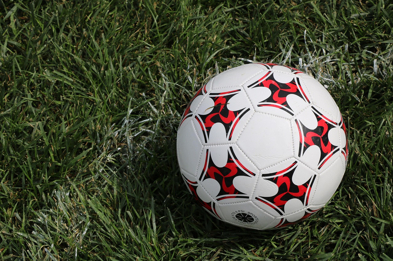 Ein weißer Fußball mit einem Muster aus schwarzen und roten geometrischen Formen liegt auf einem grünen Rasenfeld und erinnert an das Fußballfieber. Das leicht abgenutzte Gras weist weiße Markierungen auf, die einen Teil einer nahe gelegenen Feldbegrenzungslinie anzeigen. Die strukturierte Oberfläche des Balls reflektiert subtil das Sonnenlicht. Diese Beschreibung wurde mit der FootballR KI automatisch generiert.