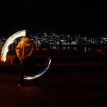 Eine Person führt nachts an einem Ufer Feuerwirbel vor, im Hintergrund sind die Lichter der Stadt und die Space Needle zu sehen. Sie trägt einen Kapuzenpullover und verschwimmt durch die Bewegung, sodass Lichtbögen entstehen, wenn sie die Feuerpoi schwingt. Diese Beschreibung wurde mit der FootballR KI automatisch generiert.