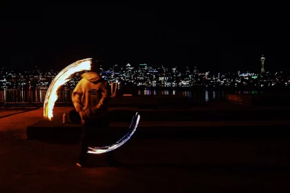 Eine Person führt nachts an einem Ufer Feuerwirbel vor, im Hintergrund sind die Lichter der Stadt und die Space Needle zu sehen. Sie trägt einen Kapuzenpullover und verschwimmt durch die Bewegung, sodass Lichtbögen entstehen, wenn sie die Feuerpoi schwingt. Diese Beschreibung wurde mit der FootballR KI automatisch generiert.