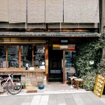 Straßenansicht eines malerischen Cafés mit Holzdekor und einladendem Eingang. Draußen steht neben einem Menüständer ein rosa Fahrrad. Das Geschäft verfügt über große, rustikale Fenster und viel Grün, was eine gemütliche Atmosphäre schafft. Auf einer kleinen Tafel werden die Tagesgerichte angezeigt. Diese Beschreibung wurde mit der FootballR KI automatisch generiert.