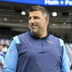 Mike Vrabel steht in einem blau-marineblauen Sportjackett drinnen und blickt nach links. Auf der Brust des Jacketts ist ein Logo zu sehen. Hinter ihm füllen verschwommene Zuschauer ein Stadion, und oben auf einem großen Bildschirm wird Text angezeigt. Die Kulisse scheint eine geschäftige Sportveranstaltung zu sein. Diese Beschreibung wurde mit der FootballR KI automatisch generiert.