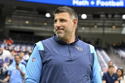 Mike Vrabel steht in einem blau-marineblauen Sportjackett drinnen und blickt nach links. Auf der Brust des Jacketts ist ein Logo zu sehen. Hinter ihm füllen verschwommene Zuschauer ein Stadion, und oben auf einem großen Bildschirm wird Text angezeigt. Die Kulisse scheint eine geschäftige Sportveranstaltung zu sein. Diese Beschreibung wurde mit der FootballR KI automatisch generiert.