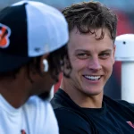 Woche 13 - Joe Burrow und Ja'Marr Chase sitzen im Freien. Die Person rechts lächelt und trägt ein schwarzes T-Shirt mit einem kleinen Logo. Die Person links trägt eine Bengals-Kappe, wahrscheinlich zu Ehren von Joe Burrow, und einen Kapuzenpullover und blickt in Richtung der lächelnden Person. Der Hintergrund ist unscharf und konzentriert sich auf ihre Interaktion. Diese Beschreibung wurde mit der FootballR KI automatisch generiert.