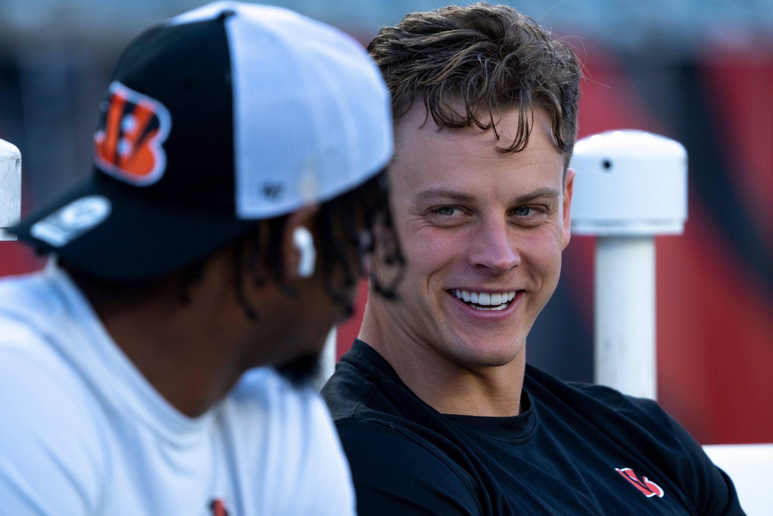 Joe Burrow und Ja'Marr Chase sitzen im Freien. Die Person rechts lächelt und trägt ein schwarzes T-Shirt mit einem kleinen Logo. Die Person links trägt eine Bengals-Kappe, wahrscheinlich zu Ehren von Joe Burrow, und einen Kapuzenpullover und blickt in Richtung der lächelnden Person. Der Hintergrund ist unscharf und konzentriert sich auf ihre Interaktion. Diese Beschreibung wurde mit der FootballR KI automatisch generiert.