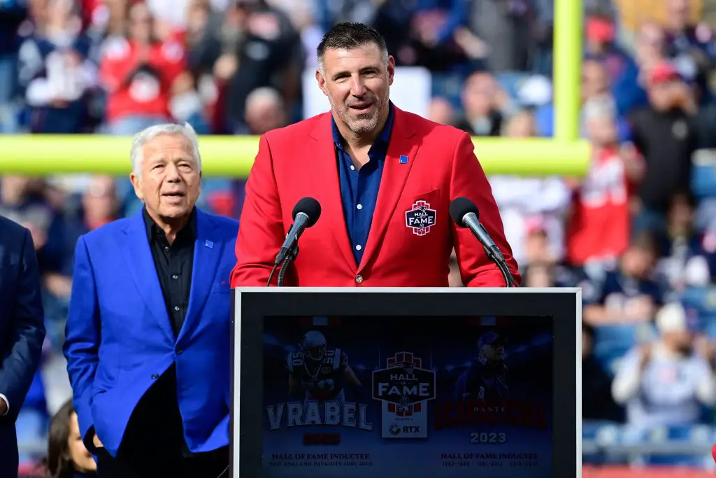 Mike Vrabel in einem leuchtend roten Blazer, geschmückt mit einem Hall of Fame-Abzeichen, spricht an einem Podium mit der Aufschrift „Vrabèl Hall of Fame Inductee 2023“. Mike Vrabel, bekannt für seine Zeit bei den New England Patriots, steht neben einem anderen Mann in Dunkelblau. Eine verschwommene Menschenmenge und Football-Torpfosten füllen den Hintergrund. Diese Beschreibung wurde mit der FootballR KI automatisch generiert.