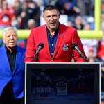 Week 16 - Mike Vrabel in einem leuchtend roten Blazer, geschmückt mit einem Hall of Fame-Abzeichen, spricht an einem Podium mit der Aufschrift „Vrabèl Hall of Fame Inductee 2023“. Mike Vrabel, bekannt für seine Zeit bei den New England Patriots, steht neben einem anderen Mann in Dunkelblau. Eine verschwommene Menschenmenge und Football-Torpfosten füllen den Hintergrund. Diese Beschreibung wurde mit der FootballR KI automatisch generiert.