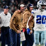 Footballtrainer Brian Schottenheimer, in einem hellbraunen Kapuzenpulli und einer schwarzen Kappe gekleidet, ruft leidenschaftlich von der Seitenlinie. Er hält eine Spielkarte in der Hand und steht neben einem Spieler in einer silber-blauen Uniform. Im Hintergrund sind weitere Trainer und Teammitglieder zu sehen. Diese Beschreibung wurde mit der FootballR KI automatisch generiert.