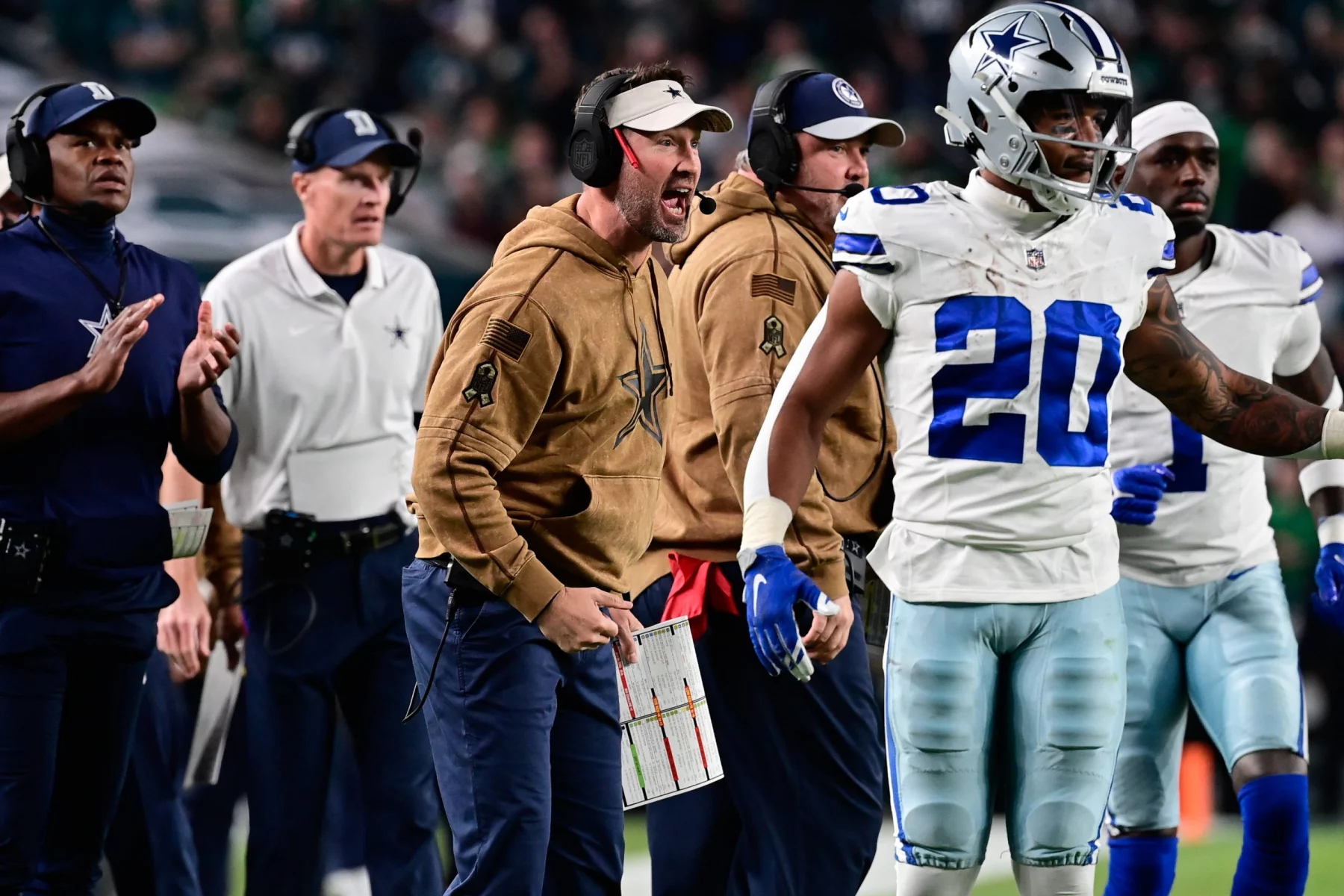 Footballtrainer Brian Schottenheimer, in einem hellbraunen Kapuzenpulli und einer schwarzen Kappe gekleidet, ruft leidenschaftlich von der Seitenlinie. Er hält eine Spielkarte in der Hand und steht neben einem Spieler in einer silber-blauen Uniform. Im Hintergrund sind weitere Trainer und Teammitglieder zu sehen. Diese Beschreibung wurde mit der FootballR KI automatisch generiert.