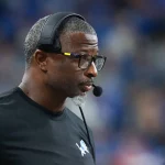 Aaron Glenn mit kurzen Haaren, Brille und Headset-Mikrofon ist konzentriert und blickt zur Seite, als würde Aaron Glenn am Spielfeldrand coachen. Er trägt ein schwarzes Shirt mit einem kleinen vogelähnlichen Logo. Der unscharfe Hintergrund lässt auf eine spannende Sportszene schließen. Diese Beschreibung wurde mit der FootballR KI automatisch generiert.