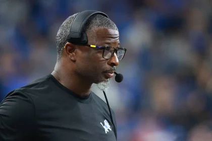 Aaron Glenn mit kurzen Haaren, Brille und Headset-Mikrofon ist konzentriert und blickt zur Seite, als würde Aaron Glenn am Spielfeldrand coachen. Er trägt ein schwarzes Shirt mit einem kleinen vogelähnlichen Logo. Der unscharfe Hintergrund lässt auf eine spannende Sportszene schließen. Diese Beschreibung wurde mit der FootballR KI automatisch generiert.