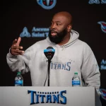 Ein Mann namens Ran Carthon steht an einem Podium mit dem Logo der Tennessee Titans und trägt einen grauen Titans-Kapuzenpulli. Er gestikuliert mit einer Hand, während zwei Wasserflaschen auf dem Podium stehen. Im Hintergrund ist das Logo der Titans neben dem Text „Ascension Saint Thomas“ deutlich zu sehen. Diese Beschreibung wurde mit der FootballR KI automatisch generiert.