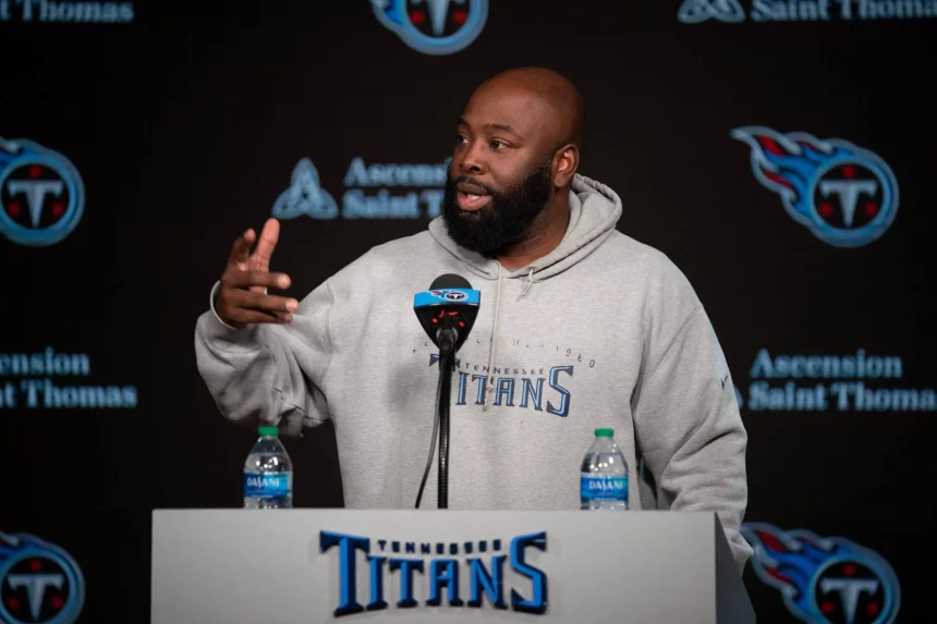 Ein Mann namens Ran Carthon steht an einem Podium mit dem Logo der Tennessee Titans und trägt einen grauen Titans-Kapuzenpulli. Er gestikuliert mit einer Hand, während zwei Wasserflaschen auf dem Podium stehen. Im Hintergrund ist das Logo der Titans neben dem Text „Ascension Saint Thomas“ deutlich zu sehen. Diese Beschreibung wurde mit der FootballR KI automatisch generiert.