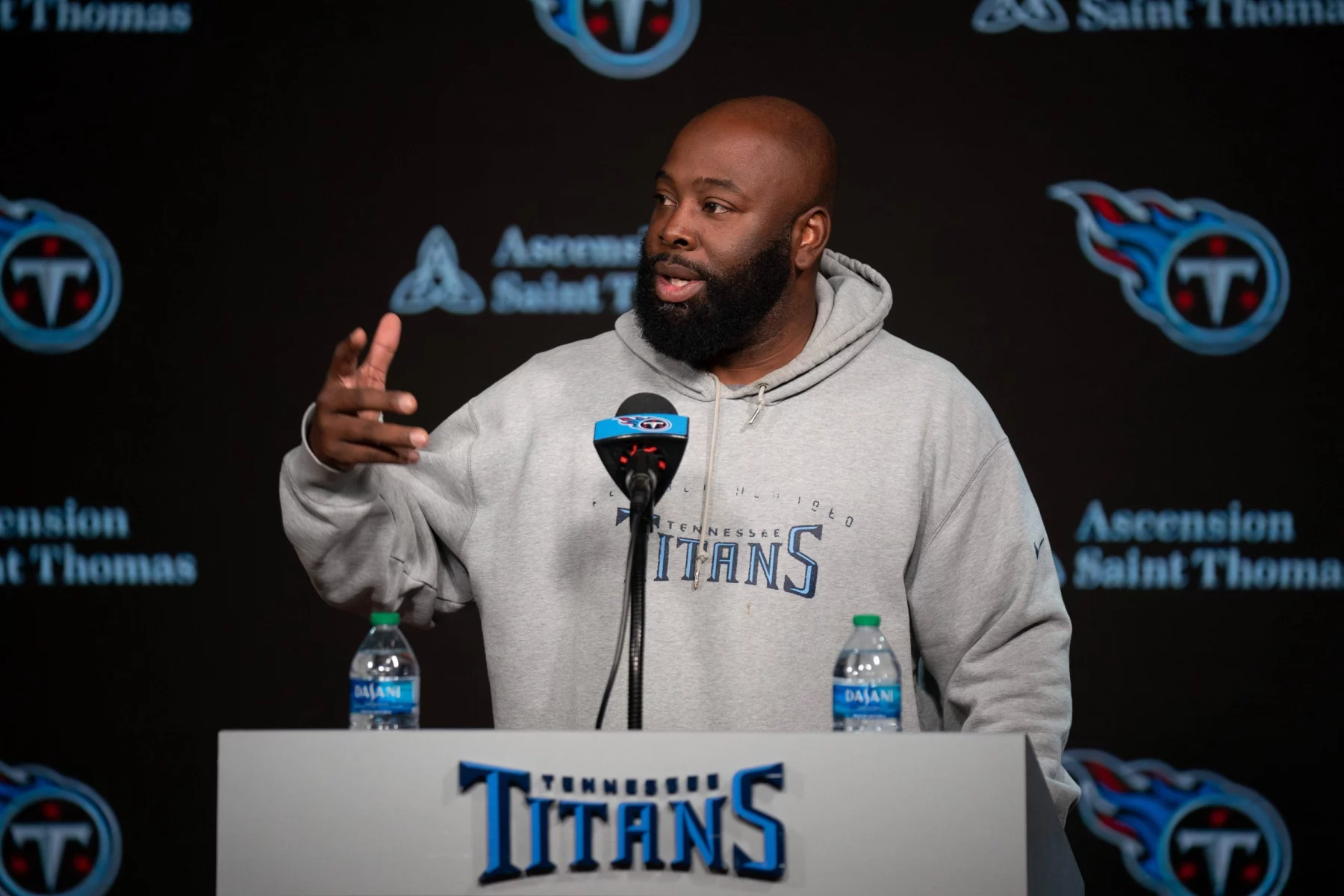 Ein Mann namens Ran Carthon steht an einem Podium mit dem Logo der Tennessee Titans und trägt einen grauen Titans-Kapuzenpulli. Er gestikuliert mit einer Hand, während zwei Wasserflaschen auf dem Podium stehen. Im Hintergrund ist das Logo der Titans neben dem Text „Ascension Saint Thomas“ deutlich zu sehen. Diese Beschreibung wurde mit der FootballR KI automatisch generiert.