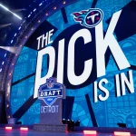 Cam Ward - NFL Draft Reihenfolge: Auf einer großen Bühne wird „The Pick Is In“ neben einem NFL Draft 2024-Logo gezeigt, wobei „Detroit“ prominent dargestellt ist. Im Hintergrund sind Tennessee Titans-Logos auf einer blauen Leinwand zu sehen, die auf die Reihenfolge des Drafts hinweisen. Helle Bühnenlichter beleuchten Menschen, die links neben einem kleinen Podium stehen. Diese Beschreibung wurde mit der FootballR KI automatisch generiert.