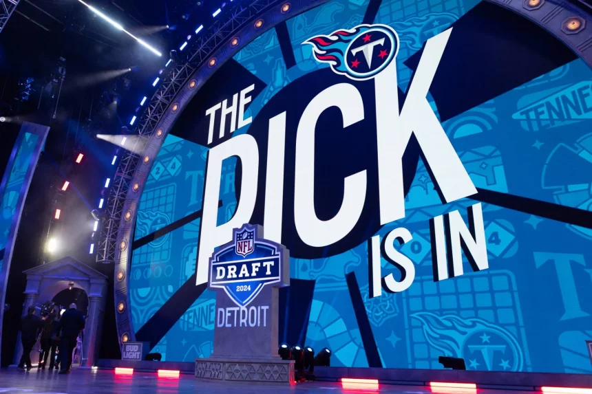 Cam Ward - NFL Draft Reihenfolge: Auf einer großen Bühne wird „The Pick Is In“ neben einem NFL Draft 2024-Logo gezeigt, wobei „Detroit“ prominent dargestellt ist. Im Hintergrund sind Tennessee Titans-Logos auf einer blauen Leinwand zu sehen, die auf die Reihenfolge des Drafts hinweisen. Helle Bühnenlichter beleuchten Menschen, die links neben einem kleinen Podium stehen. Diese Beschreibung wurde mit der FootballR KI automatisch generiert.