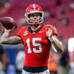 Carson Beck, der ein rotes Trikot mit der Nummer 15, schwarze Hosen und einen roten Helm trägt, bereitet sich darauf vor, auf das Feld zu werfen. Im Hintergrund sind die Zuschauer zu sehen, während er seinen rechten Arm nach hinten streckt, den Football in der Hand hält und seinen linken Arm nach vorne streckt. Diese Beschreibung wurde mit der FootballR KI automatisch generiert.