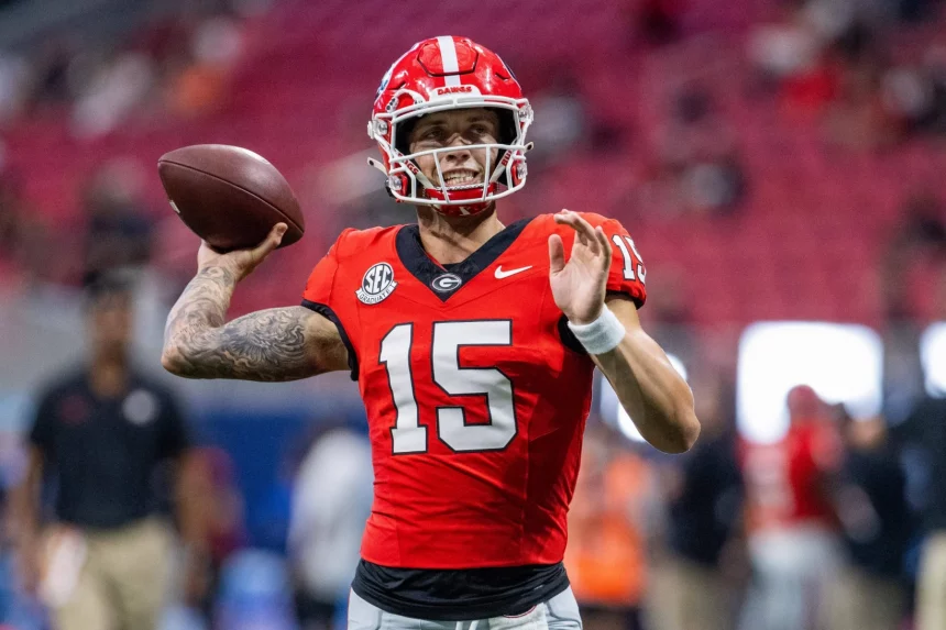 Carson Beck, der ein rotes Trikot mit der Nummer 15, schwarze Hosen und einen roten Helm trägt, bereitet sich darauf vor, auf das Feld zu werfen. Im Hintergrund sind die Zuschauer zu sehen, während er seinen rechten Arm nach hinten streckt, den Football in der Hand hält und seinen linken Arm nach vorne streckt. Diese Beschreibung wurde mit der FootballR KI automatisch generiert.