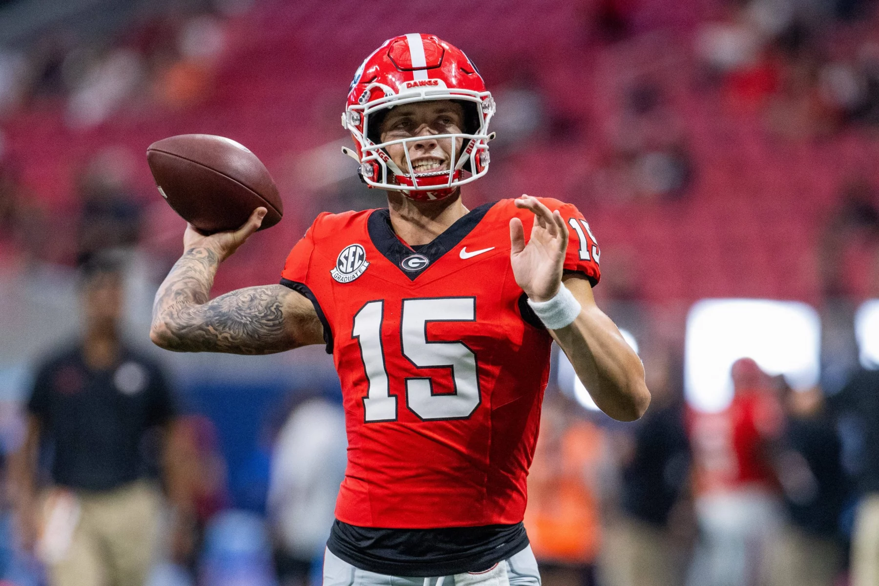 Carson Beck, der ein rotes Trikot mit der Nummer 15, schwarze Hosen und einen roten Helm trägt, bereitet sich darauf vor, auf das Feld zu werfen. Im Hintergrund sind die Zuschauer zu sehen, während er seinen rechten Arm nach hinten streckt, den Football in der Hand hält und seinen linken Arm nach vorne streckt. Diese Beschreibung wurde mit der FootballR KI automatisch generiert.
