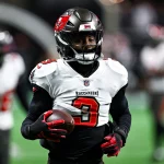 Jordan Whitehead trägt ein weiß-rotes Trikot der Tampa Bay Buccaneers mit der Nummer 3 und hält einen Football in der Hand. Mit Helm und schwarzen Handschuhen steht er unter künstlichem Licht bereit auf dem Feld und verkörpert Teamgeist, während im Hintergrund ein anderer Spieler auftaucht. Diese Beschreibung wurde mit der FootballR KI automatisch generiert.