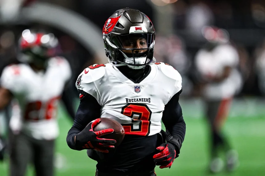 Jordan Whitehead trägt ein weiß-rotes Trikot der Tampa Bay Buccaneers mit der Nummer 3 und hält einen Football in der Hand. Mit Helm und schwarzen Handschuhen steht er unter künstlichem Licht bereit auf dem Feld und verkörpert Teamgeist, während im Hintergrund ein anderer Spieler auftaucht. Diese Beschreibung wurde mit der FootballR KI automatisch generiert.