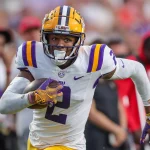 LSU-Star Kyren Lacy stürmt in seiner ikonischen Uniform mit dem Ball in der Hand nach vorne. Er trägt einen gelben Helm mit violetten Streifen und dazu passende Handschuhe. Die verschwommene Menge und die Spieler in roter Kleidung rahmen seinen konzentrierten, entschlossenen Gesichtsausdruck ein, während er über das Feld stürmt. Diese Beschreibung wurde mit der FootballR KI automatisch generiert.