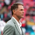 Trent Baalke, in einem hellgrauen Anzug mit weißem Hemd, steht konzentriert auf dem Fußballfeld. Sein Blick geht nach links, während der unscharfe Hintergrund Stadionsitze in Rosatönen und undeutliche Gestalten in Sportuniformen zeigt. Diese Beschreibung wurde mit der FootballR KI automatisch generiert.