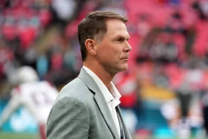 Trent Baalke, in einem hellgrauen Anzug mit weißem Hemd, steht konzentriert auf dem Fußballfeld. Sein Blick geht nach links, während der unscharfe Hintergrund Stadionsitze in Rosatönen und undeutliche Gestalten in Sportuniformen zeigt. Diese Beschreibung wurde mit der FootballR KI automatisch generiert.