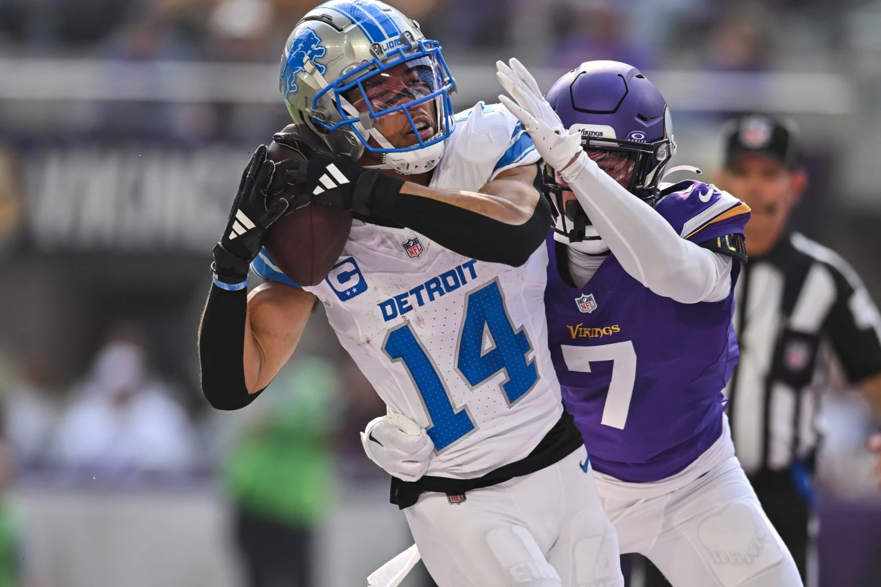Heute im TV - In Woche 18 fängt der Spieler Nr. 14 der Detroit Lions einen Football, während er von dem Spieler Nr. 7 der Minnesota Vikings eng bewacht wird. Der Spieler der Lions trägt eine weiß-blaue Uniform und einen Helm, während der Spieler der Vikings lila-weiß ist. Im Hintergrund ist ein Schiedsrichter zu sehen. Diese Beschreibung wurde mit der FootballR KI automatisch generiert.