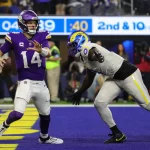 NFL Playoffs & NFL Offseason: Ein Footballspieler in violetter Uniform und mit Helmnummer 14 bereitet sich darauf vor, den Ball zu werfen, während er von einem gegnerischen Spieler in weißer Uniform und mit blau-gelbem Helm (Nummer 90) verfolgt wird. Die spannende Action spielt sich während der NFL-Playoffs auf einem blauen Feld ab, im Hintergrund ist eine Markierung für den 2. und 10. Versuch zu sehen. Diese Beschreibung wurde mit der FootballR KI automatisch generiert.